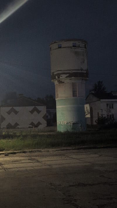 The image depicts a concrete structure, likely a water tower, at night in Gorin, Russia. The tower is cylindrical with a lighter-colored top section that appears weathered and slightly dilapidated. Below this, a band of teal or light green paint wraps around the structure, also showing signs of wear.

To the right of the tower, there is a building with a patterned facade featuring large, dark, chevron-like shapes arranged in a symmetrical pattern. This building also appears to be lit from the side, casting light onto the adjacent grassy area and a paved surface. The paved surface, likely a road or pathway, has tire tracks and appears damp, suggesting recent rain or dew.

The overall scene is dimly lit by artificial light sources, creating shadows and highlighting the textures of the concrete and pavement. The sky is dark, indicating it is nighttime. The environment appears to be an urban or semi-urban setting, with hints of buildings and vegetation. There is no visible text in the image. The overall mood is somewhat stark and quiet, fitting for a nighttime industrial or infrastructure scene.