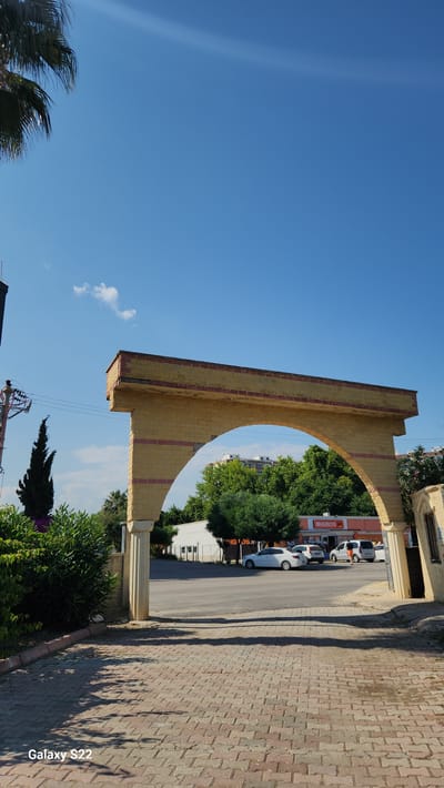 The image depicts an outdoor scene in Erdemli, Turkey, on a clear, sunny day. A large, yellow-orange brick or stone archway, adorned with horizontal reddish-pink stripes, dominates the central midground. The archway's left support features a classical column, while its right support connects to a similarly constructed wall section with a window opening.
In the foreground, a pathway paved with reddish-brown interlocking pavers extends towards the archway, exhibiting distinct shadows cast from the left, suggesting a sun position to the right. To the far left, a portion of a palm tree frond is visible against the bright blue sky. Below it, a dark utility pole with overhead wires and a tall, slender coniferous tree stand. Dense green bushes are visible at the lower left.
Through the archway, an asphalt road is visible, with at least three parked vehicles: a white sedan, a light-colored hatchback, and a white van, all facing away from the viewer. Behind the vehicles, a building with a prominent orange "MIGROS" supermarket sign is clearly identifiable. Beyond the supermarket and the road, abundant green trees partially obscure taller, multi-story residential buildings in the distance. The sky above is a uniform clear blue, with a single small white cloud visible to the left of the archway's apex. A faint sun ray or lens flare is observable in the upper left quadrant of the sky. No individuals are explicitly visible within the frame. The bottom left corner of the image contains a "Galaxy S22" watermark.