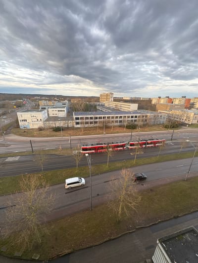 The image presents an elevated, wide-angle view of an urban environment in Halle (Saale), Germany, specifically depicting the area around Heideallee and Weinberg Campus. The sky is heavily overcast with a uniform layer of dense, grey stratocumulus clouds, casting a diffused light over the scene. Hints of brighter, diffused light are visible on the distant horizon, suggesting either early morning or late afternoon.

From an elevated vantage point, evidenced by the partially visible dark, flat roof edge in the bottom right corner, the scene looks across a wide, multi-lane asphalt road that traverses horizontally through the mid-ground. This road features a central reservation containing double tram tracks. A three-segment, articulated red and white tram is visible on these tracks, oriented towards the right side of the frame. On the road lanes, a white van is present in the lane closer to the viewer, moving right, and a dark-colored sedan is further ahead, also moving right. Both the road's verges and the central reservation are lined with bare deciduous trees, indicating a dormant vegetative season, and sparsely covered with brownish-green grass. Gray street light poles are interspersed along the road.

Beyond the road and tram tracks, the mid-ground features a prominent, long, multi-story building with a light-colored facade and numerous rectangular windows. Its flat roof is extensively covered with solar panels. To the left of this building, another smaller, light-colored building, exhibiting some graffiti on its exterior, is visible near a road intersection.

The background unfolds into an expansive urban landscape comprised of various buildings, including taller multi-story structures, residential blocks with varied colored facades, and smaller commercial or institutional units. A line of distant, bare trees marks the horizon, suggesting the city's edge. No human individuals are discernible within the visible area.