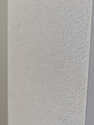 The image shows a close-up, angled view of a textured white wall. The texture is rough and uneven, with small, irregular bumps and ridges creating a pebbled appearance. To the left of the textured wall, a smoother, lighter-colored surface is visible, suggesting a corner or an adjacent wall. The lighting appears to be soft and diffuse, casting subtle shadows that highlight the texture of the wall. There are no people, objects, or actions depicted in the image. The provided context about the location in Settimo Torinese, Italy, is not visually evident from the image itself.