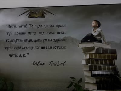 The image is a photograph of a mural or artwork that features a quote by Ivan Vazov, a prominent Bulgarian writer. The artwork is located in Dalgopol, Bulgaria. The quote, written in Bulgarian, is: "Чети, чети! То чудо днеска прави; туй дребно нещо под това небе, то мъртви буди, дава ум на здрави, туй второ слънце Бог ни сам остави: чети: а, б.” This translates to: "Read, read! Today it performs a miracle; this small thing under this sky, it raises the dead, gives sense to the healthy, this second sun God himself left us: read: a, b."
The artwork depicts a young boy sitting on a stack of large, old books, looking thoughtfully into the distance. The background is a dark, cloudy sky, creating a dramatic and somewhat ethereal atmosphere. Above the quote, there is an image of an open book with rays of light emanating from it, symbolizing knowledge and enlightenment. The signature "Иван Вазов" (Ivan Vazov) is visible in cursive script below the quote.
The overall impression is one of education, wisdom, and the power of reading. The location in Dalgopol, Bulgaria, suggests this artwork is a cultural installation intended to promote literacy and honor the legacy of Ivan Vazov.