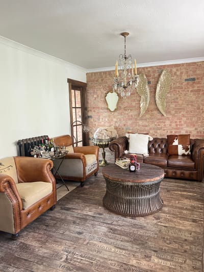 This media file captures an interior scene featuring a collection of furniture and decorative items arranged in a living room or lounge area. The room is characterized by exposed brick walls and a rustic wooden floor with visible text and dates printed on it, suggesting a vintage or industrial aesthetic.
Dominating the scene are several pieces of brown leather furniture, including a large tufted sofa with cowhide and plain accent pillows, and two accent chairs. A round wooden coffee table with a metal-ribbed base sits in front of the sofa. A bar cart laden with bottles and glasses is positioned near one of the accent chairs, indicating a possible social gathering or relaxation setting.
Above the sofa, two large, golden wing-shaped wall decorations are mounted on the brick wall. A crystal chandelier hangs from the ceiling, casting a warm light on the space. A small ornate mirror is also visible on the wall.
The overall impression is one of a stylish, somewhat eclectic lounge with a blend of rustic and luxurious elements. The lighting suggests it might be evening or an overcast day, as natural light is not prominently featured. The visible text on the floor appears to be a repeating pattern of words and numbers, possibly related to shipping manifests or vintage labels. There are no people visible in the image.