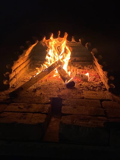 An indoor fireplace is lit, consuming logs within a stone structure with a decorative, notched front. The scene is predominantly dark, highlighting the bright orange and yellow flames. The fireplace walls are constructed of rough-hewn stones. Konstantinovo, Bulgaria, is not discernible from the image.