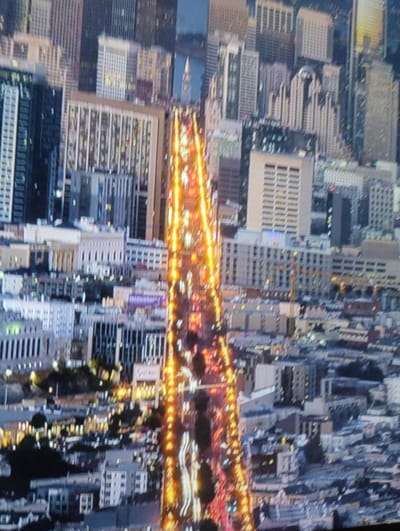 The image is a blurry, high-angle aerial view of a city at night, with a focus on a brightly lit street lined with buildings. The street appears to be a major thoroughfare, with numerous car headlights and taillights creating streaks of light. The surrounding buildings are tall and modern, with many windows illuminated. The overall impression is of a bustling urban environment. The question asks to include the location of Rechytsa, Belarus. However, this image does not appear to be of Rechytsa, Belarus. It is highly likely that this is an image of a large North American city, possibly San Francisco due to the architecture and the bright lights of a major street. There are no people visible in the image, only the lights of vehicles and buildings. The image is taken from a high vantage point, suggesting it might be from a drone or a very tall building.