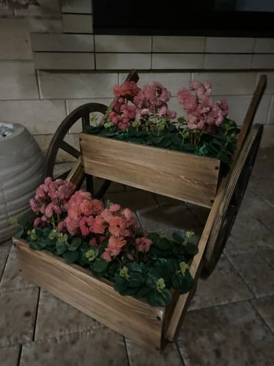 This media file shows a wooden garden cart filled with artificial pink flowers and greenery. The cart has two tiers, with the flowers and foliage cascading over the sides. The cart is positioned outdoors on a tiled surface. In the background, there appears to be a wall made of rectangular tiles and a darker, possibly wooden, structure. The lighting suggests it could be evening or nighttime, with artificial light sources illuminating the scene. The overall impression is one of decorative landscaping, possibly in a yard or on a patio. There are no people visible in the image. The image is oriented vertically, with the cart appearing at an angle.