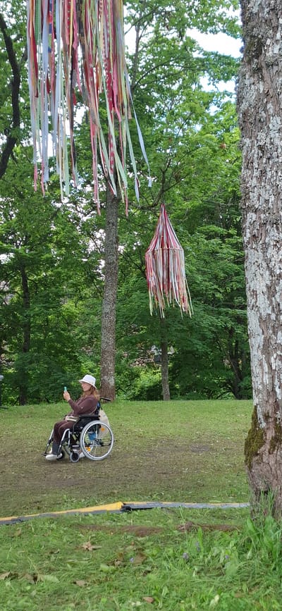 A medium shot shows a woman in a wheelchair looking at her phone, sitting on a grassy hill with trees in the background. She is wearing a white hat and a brown hooded sweatshirt. The wheelchair has large rear wheels and smaller front wheels. Colorful ribbons are hanging from trees in the foreground, creating a festive atmosphere. The overall setting appears to be an outdoor event or park. The image was taken in Viljandi, Estonia.