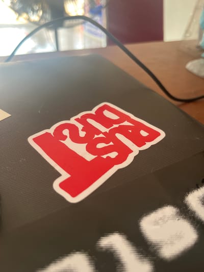 The media file shows a close-up, angled shot of a laptop with a red and white sticker on its surface. The sticker has bold, stylized text that appears to read "BRUT." The background of the laptop lid is a textured black. To the left of the laptop, a black cable is visible, snaking across a wooden surface. In the blurred background, parts of what looks like a desk or table are visible, with some indistinct objects and a hint of blue fabric. The lighting suggests an indoor setting, possibly a room during the daytime. The image focuses on the sticker and the immediate surrounding area of the laptop, providing no specific cues about the time of day, weather, or a precise location beyond what might be inferred from the general scene. No individuals are depicted in the image.