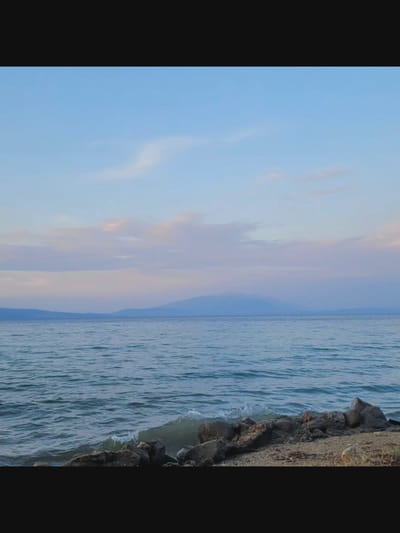 The image depicts a serene coastal landscape under a soft, pastel sky. The foreground features a rocky shoreline with small waves gently lapping at the stones. Beyond the immediate shore, a vast expanse of calm water stretches towards a distant, hazy mountain range. The sky is a gradient of pale blue and soft pinks and purples, suggesting either dawn or dusk. There are no people or man-made structures visible in the scene. The overall impression is one of natural tranquility. The location is identified as Bielawska Wieś, Poland, a detail that does not provide specific visual cues within the image itself.