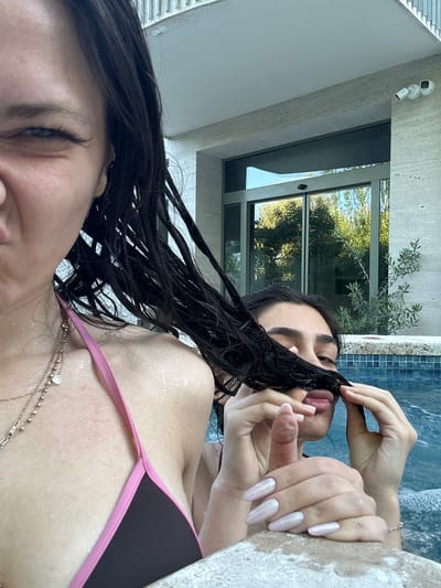 This media file shows two young women in a hot tub, likely outdoors given the lush green trees visible through a large window in the background. One woman, in the foreground and to the left, is looking down with her eyes partially closed, her dark, wet hair falling around her face and shoulders. She is wearing a dark bikini top with pink trim and a delicate necklace with a heart-shaped pendant. The other woman is further back in the hot tub, her face partially obscured as she pulls a lock of her own dark, wet hair towards her mouth. Her hands are visible, one reaching for her hair and the other resting near her face. She has long, light pink manicured nails. The water in the hot tub is a vibrant blue, with a tiled edge. The surrounding architecture suggests a modern building with stone accents and a large window. Two security cameras are mounted on the exterior wall near the window. The scene appears to be from daytime, with bright natural light illuminating the area. The overall mood seems relaxed and intimate.