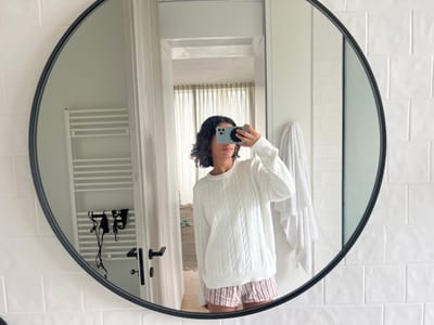The image presents a mirror selfie taken in a bathroom located in Vila Nova de Cerveira, Portugal. A circular mirror with a black frame reflects a person, whose face is partially obscured by a light-colored smartphone held horizontally to take the photograph. The individual wears a white, cable-knit long-sleeved sweater and light-colored, striped shorts. Visible in the reflection behind the subject is a white tiled wall. To the left, a white, horizontally-barred towel radiator is mounted, with a black hairdryer and its cord hanging below it. To the right, a white towel is draped over a fixture. An open doorway in the background reveals an adjacent room with light-colored flooring and a window featuring sheer curtains.