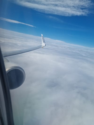 The image shows a view from an airplane window. The wing of the plane is visible, with the Ryanair logo on its tip. An engine is visible at the right side of the image, with the foreground partially showing the interior of the plane. Outside the window, there is a view of clouds and blue sky. The location, Breze, Bulgaria, cannot be determined from the visual information.