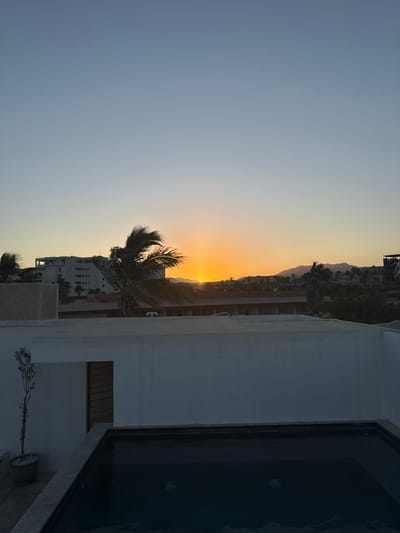 The image presents an elevated, static view of San José del Cabo, Mexico, captured during either sunrise or sunset. In the immediate foreground, a dark-tiled, rectangular plunge pool with light-colored coping is situated on a rooftop patio. Adjacent to the pool on the left, a white-painted concrete structure incorporates a vertical wooden slatted door and a small potted plant. The midground features several palm trees with fronds appearing to be wind-blown, set against a backdrop of various light-colored buildings, one of which is a multi-story structure on the left with visible exposed rebar on its upper level, suggesting ongoing construction or an unfinished state. A reddish-brown rooftop is visible below these structures. The horizon is dominated by a brilliant orange and yellow sun, positioned centrally, which casts a bright glow and silhouettes distant mountain ranges and hills. The sky transitions from intense orange and yellow near the horizon to a clear, pale blue at the top of the frame. No individuals, animals, or active subjects are present in the scene.