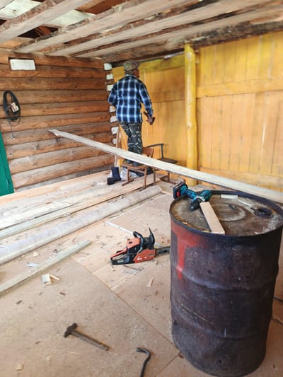 The image depicts the interior of a rustic, unfinished wooden structure, likely a barn or workshop, with natural light illuminating the scene. The walls are constructed from horizontal logs, and the ceiling consists of exposed wooden beams. In the center of the frame, a person, wearing a plaid shirt, camouflage pants, and a cap, is seen from the back, walking away from the camera. They appear to be involved in some construction or renovation work. Scattered on the wooden floor are various tools and materials, including a chainsaw, a hammer, a drill, a large metal barrel, and numerous wooden planks. Some of the planks are long and appear to be newly cut, while others are smaller pieces. The overall impression is one of ongoing manual labor in a rural setting, possibly in the village of Islambaevo, Russia, given the architectural style and rural elements.