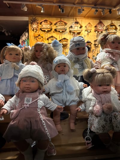 In a brightly lit, indoor market stall in Mayrhofen, Austria, a collection of lifelike baby dolls is displayed on wooden shelves. The dolls are dressed in winter-themed outfits, including knitted hats, scarves, and jackets, many adorned with pom-poms and bows. Some dolls are accessorized with pacifiers and chains. In the background, a wall of cuckoo clocks adds to the festive atmosphere. The dolls are arranged in rows, with some closer to the viewer than others. The lighting is warm and artificial, casting shadows on the dolls and the wooden surfaces.
