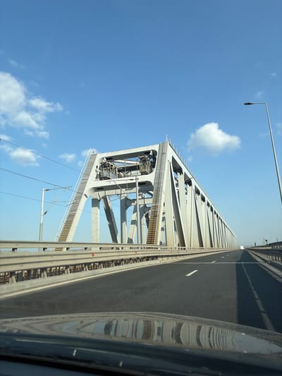 The image displays the Cernavodă Bridge, situated near Stelnica, Ialomița County, Romania. The scene is captured from a forward-facing, low-angle perspective within a vehicle traveling on the bridge's roadway. A large, light-grey steel truss bridge structure dominates the upper half of the frame, featuring intricate geometric bracing and elevated maintenance access stairs on its primary supports. The bridge accommodates both vehicular traffic on the left and a railway line parallel to it on the right. The asphalt roadway in the foreground is black with white lane markings, including a solid line on the left edge and a dashed center line. Metal guardrails with red reflective markers separate the road from the railway and the bridge's edge. Overhead electrical catenary lines for the railway are visible on the left side of the bridge structure. A tall, dark street lamp post stands on the far right. In the distant background on the roadway, a small white vehicle, possibly a truck or van, is visible. The sky is clear and bright blue with scattered white cumulus clouds. Reflections of the vehicle's dashboard are present across the bottom of the image. No people are visible.