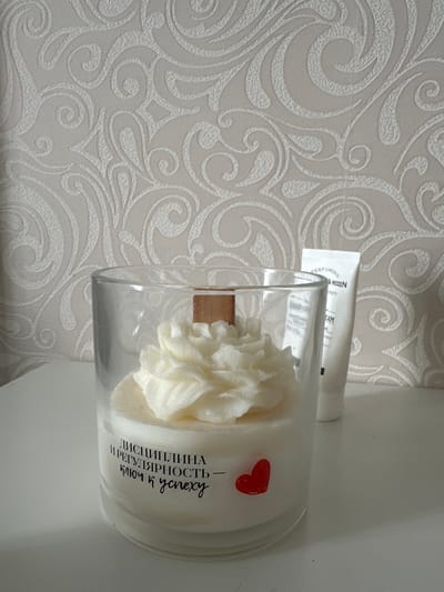 An indoor shot captures a candle in a glass jar with a decorative cream topping and a wooden wick, placed on a white surface. To the right of the candle, a white tube with text, possibly hand cream, stands upright. The background features textured wallpaper with a swirling pattern. The candle has text and a small red heart decal on its side. The scene does not provide any information about the city of Malye Ust'ya, Russia.