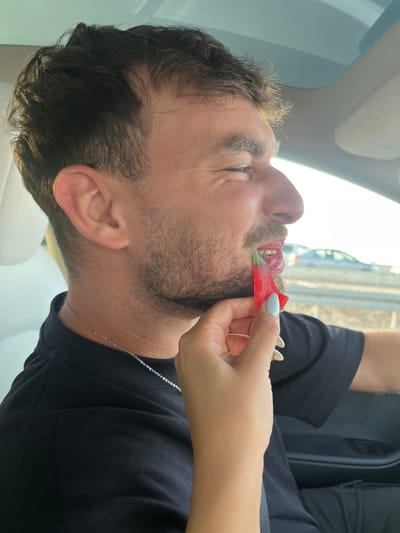 A male individual with dark, wavy hair and stubble is depicted from the right side, seated in the driver's position of a vehicle in Kfar Qasim, Israel. He wears a black t-shirt and a silver chain. His right eye is squinted, and his mouth is open with his tongue extended, displaying green residue. A female's right hand, with light blue manicured nails, holds a red, sugar-coated, chili-pepper-shaped gummy candy with a green stem, which is partially inserted into the male's mouth. The vehicle interior features a light-colored headliner and dark accents. Through the window, an exterior view of an asphalt road, a guardrail, and blurred vehicles in the distance is visible under daylight conditions.