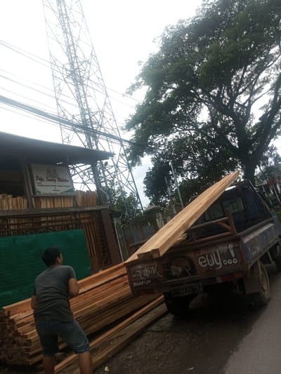 In the city of Sarwahi Mal., India, an outdoor scene depicts a man engaged in handling lumber next to a blue pickup truck. A single male figure is visible from the back, wearing a dark short-sleeved shirt and blue shorts, positioned near a large stack of light-colored wooden planks on the ground. A blue pickup truck, branded "SUZUKI" on the tailgate, is parked on a wet, dark surface, with graffiti including "euy..." and other markings. Its license plate displays partially visible characters "0239 YJ". Several long wooden planks are loaded onto the truck's bed, with one prominent plank extending diagonally upwards and rearwards. To the left, a building features a wooden facade and a sign reading "P.T.K. Sarwahi Walaka" with additional text and contact information. Below the sign, a green panel or tarp is visible. A tall, metal lattice communication tower with numerous power lines dominates the background against an overcast sky. Large, dense green trees are visible to the right of the tower. The ground appears wet, suggesting damp conditions.