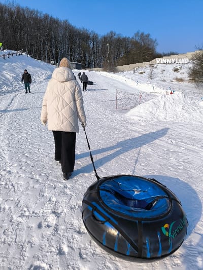 This image depicts a winter scene at Vinny Land in Prybuzke, Ukraine. The foreground shows an individual from behind, wearing a beige quilted jacket, a light-colored knitted beanie, black pants, and dark boots, walking uphill on a snow-covered slope. This person is pulling a black and blue inflatable snow tube, featuring a "Vinny Land" logo, via a black strap. Long shadows are cast on the snow, indicating a low sun angle.
Further up the slope, several other individuals are visible; one person on the left wears a dark jacket and green pants, while two others in the distance also walk uphill, one of whom pulls a dark snow tube. The slope is groomed with visible tracks in the snow, suggesting a tubing or sledding path. To the left, wooden fence posts line the path, and a small building and a light pole are discernible on the hillside. To the right, a large white sign spells out "VINNYLAND" against a snow-covered embankment. Sections of snow netting/fencing are also visible on the right side of the slope. The background features a dense line of bare deciduous trees and some evergreen foliage under a clear blue sky.