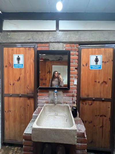 The image depicts a rustic, outdoor restroom area with two wooden doors, one marked with a male symbol and the text "SANITARIO CABALLEROS" and the other with a female symbol and the text "SANITARIO DAMAS." A stone sink with a faucet is situated between the doors, in front of a brick wall. A black bottle of soap is on the counter to the right of the sink. A young woman, holding a phone, is reflected in a mirror above the sink, taking a selfie. The overall environment appears to be an outdoor space with a natural, somewhat unfinished aesthetic. While the prompt mentions Chipilo de Francisco Javier Mina, Mexico, there are no specific visual cues in the image to confirm this location.