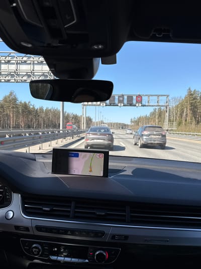 The image shows the view from inside a car driving on a highway.  The car is following other vehicles, with trees visible on either side of the road. Speed limit signs of 110 km/h are visible. Inside the car, the dashboard is visible, including the air vents, climate control panel, a screen displaying a map, and the rearview mirror. The map indicates the vehicle is approximately 34 km away from the destination. The text "КАД" can also be seen on the navigation system. Based on the text of CAD (КАД) and the speed limits, the location is likely Saint Petersburg Automobile Ring Road around Lomonosov, Russia.