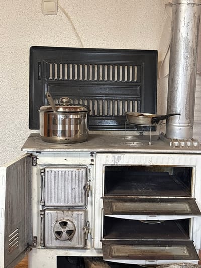 The scene, located in Hliabovo, Bulgaria, features an indoor view centered on a white, enameled cast-iron wood-burning stove positioned against a light-colored, textured wall. On the upper left section of the wall, a beige electrical switch or outlet is visible with a thin, light-colored wire extending downwards. A large, black, rectangular cast-iron grate, composed of two horizontal sections with multiple vertical slits, is mounted on the wall behind the stove.
The stove's top surface, marked with use and patina, holds two cooking items. On the left, a stainless steel pot with a fitted lid contains a metal spoon. To its right, a dark frying pan with a black handle rests on a silver-colored wire trivet, positioned over one of the stove's circular burner openings. The pan contains dark residue. A vertical, worn metallic stovepipe ascends from the right side of the stove.
The front of the stove displays multiple compartments. On the left, two closed, silver-colored metal doors are present; the lower door features a circular, three-bladed vent design. To the right, two oven or warming compartments are exposed by their respective open, downward-tilting doors, revealing dark interiors. An additional white, enameled door on the far left side of the stove is open, exposing a ribbed interior surface.