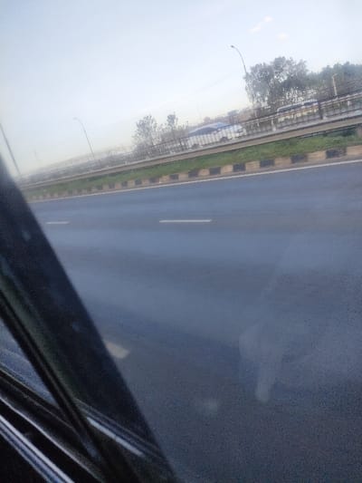 The image depicts a multi-lane highway section in Murera ward, Kenya, viewed from within a vehicle. A dark, angled structural element, likely a window frame, occupies the lower left quadrant. The primary road surface is dark asphalt, appearing damp or reflective, with two distinct white dashed lane markings. To the right, a raised median strip with green grass is visible, edged by curbs painted with alternating black and light-colored blocks. Beyond the median, an elevated highway or bridge structure runs parallel, featuring a metal barrier. A white van or large car is identifiable moving on this elevated structure, accompanied by less distinct vehicles. Sparse trees and multiple streetlights line the elevated structure and the main road. The sky is bright, hazy, and overcast.