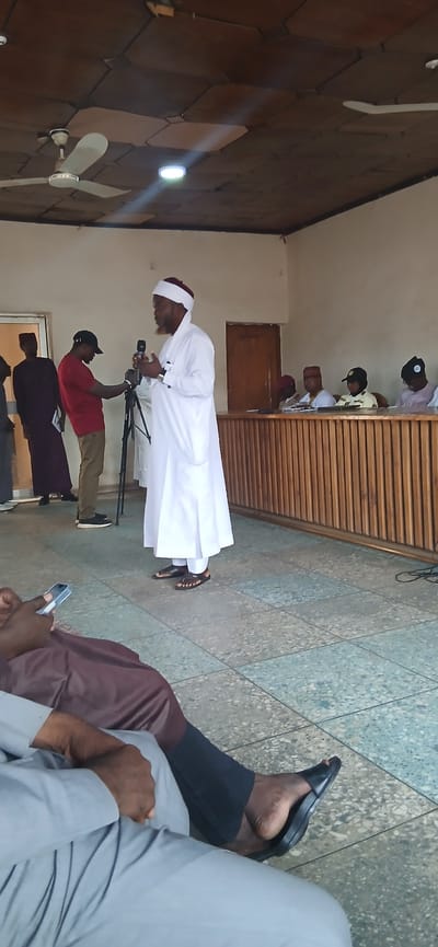 In a room with tiled floors, a group of people are gathered. The scene appears to be indoors, with a ceiling fan and wooden slatted walls visible. The overall lighting suggests it could be daytime.

The main subject is a person in the foreground wearing a white robe and a maroon turban, bending over a microphone. This person is facing towards the right side of the frame and appears to be speaking or leading an event. Behind this person, another individual in a red shirt and a black cap is holding a camera on a tripod, suggesting that the event is being filmed or documented. Further back, several other individuals are seated or standing, some wearing similar turbans and traditional attire. A wooden door is visible on the wall.

The people in the image appear to be attentive to the person speaking. The presence of a microphone and camera indicates a formal or organized gathering, possibly a religious service, a lecture, or a public address. The attire of the individuals, particularly the turbans and robes, suggests a cultural or religious context. The setting, with its simple furnishings and tiled floor, is functional and not elaborately decorated.

The provided location context of Tundun Wada West, Nigeria, further supports the likelihood of a cultural or religious event related to the community. There is no visible text in the image.