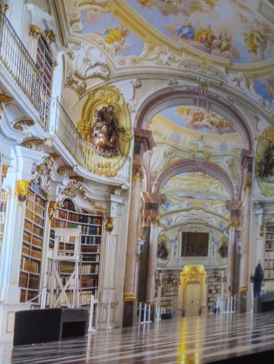 The image depicts the interior of a grand, ornate library, likely a historical or significant landmark. The architecture is Baroque or Rococo, characterized by elaborate gold ornamentation, frescoes on the ceiling, and towering bookshelves filled with leather-bound volumes. A prominent feature is a mezzanine level with a decorative railing, accessed by a staircase. In the foreground, a reflective floor with a striped pattern is visible. There are several decorative statues and elaborate architectural details throughout the space. One person, a man wearing a dark jacket and pants, is standing near the right side of the image, appearing to observe something. The overall impression is one of opulence and scholarly pursuit. The location is identified as the city of Rechytsa, Belarus.