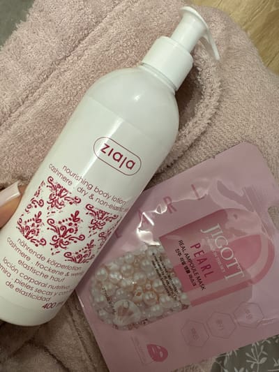 This is a close-up, top-down shot of beauty products placed on a textured, light pink fabric, possibly a robe or towel. To the left is a white pump bottle of Ziaja nourishing body lotion, with a red logo and text in multiple languages, including English "cashmere. dry & non-elastic skin" and German "nährende körperlotion cashmere. trockene & wenig elastische haut". The bottle states it contains "400 ml". To the right is a pink, rectangular packet for JIGOTT REAL AMPOULE MASK PEARL. The packet has text in English and Korean, and appears to contain a mask with small pearl-like beads. The scene is indoors, with a wooden floor visible in the upper right corner. There is no indication of the location being in Momchilovtsi, Bulgaria.