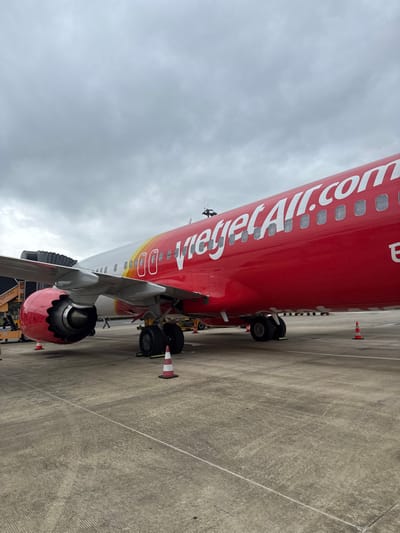 An image taken at an airport in Hòa Cường Ward, Vietnam, depicts a Vietjet Air passenger aircraft parked on a concrete tarmac under an overcast sky. The aircraft's fuselage is primarily red with "VietjetAir.com" in white lettering and a gradient of yellow, red, and white near the nose section. Multiple oval passenger windows are visible along the fuselage. The left engine, painted red with a metallic intake, is visible under the left wing. The main landing gear, featuring multiple wheels, is deployed beneath the fuselage and wing. A black accordion-style jet bridge is connected to the forward left door of the aircraft. Several orange and white traffic cones mark areas on the tarmac near the landing gear. Partially visible yellow ground service equipment is present under the right wing area. A single individual is discernible walking in the distance under the aircraft's wing. The concrete tarmac shows signs of wear and faint markings.
