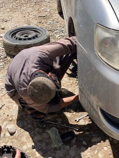The image depicts an outdoor scene in کوکران, Afghanistan, under direct sunlight. A male individual, wearing a dark brown *shalwar kameez*, dark sandals, and a decorated traditional cap, is crouched at the front driver's side of a silver sedan. The vehicle, appearing dusty, is elevated by a jack, with its front wheel removed. The man's right hand is positioned within the exposed wheel well, indicating mechanical work. A black steel wheel with a mounted tire lies on the rocky, uneven dirt ground to the man's left. Scattered on the ground near his feet are tools including a lug wrench, an open-ended wrench, and a small metal part, alongside a green plastic bag and a dark rectangular object. Strong shadows are cast on the unpaved, gravelly ground.