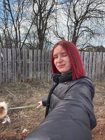 A woman with red hair, wearing a black puffer jacket, is holding a green leash attached to a small, light-colored dog. They are in an outdoor area in Заболотово, Russia, where dry grass and bare ground are visible. A wooden fence and bare tree branches with a cloudy sky in the background are present.
