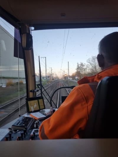 The image depicts a person wearing an orange jacket inside the cabin of a train, possibly a locomotive. The perspective is from the driver's seat, looking out onto a railway track extending into the distance. Power lines and masts are visible above the tracks, and buildings and trees are on either side. Inside the cabin, a tablet is mounted on the dashboard, displaying maps and data. Various controls and instruments are also visible. The location is in Rheinstetten, Germany, based on elements consistent with the area's railway infrastructure and architecture. The scene suggests the train is either starting, stopping, or moving slowly through the outskirts of a town.