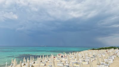 The image depicts a beach with rows of closed white beach umbrellas and lounge chairs on a sandy shore. The sea is a clear turquoise color, with some shallow areas showing the seabed. The sky is cloudy with a dark, stormy area in the distance, suggesting impending rain. Several people are visible on the beach, some sitting or lying on lounge chairs. In the far distance, there are some structures and wind turbines. This scene is characteristic of a beach resort area, likely within or near the city of Taranto, Italy, known for its coastline and beaches.