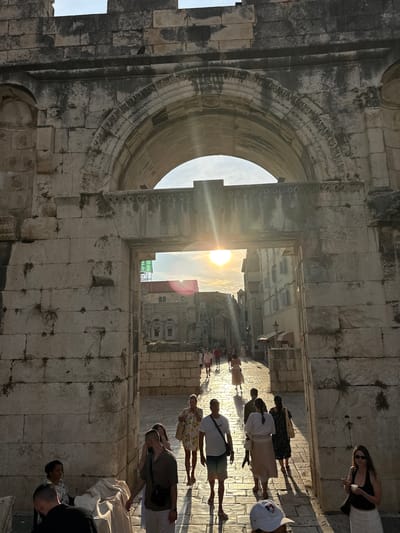 The image depicts a group of people walking through an ancient stone archway, likely in Split, Croatia, given the architectural style resembling Diocletian's Palace. The sun is setting, casting long shadows and creating a warm, golden light that streams through the archway. The sky is visible through the arch, with some clouds and lens flare from the sun. The stone structures are weathered and detailed, showing signs of age and possibly some vegetation growing in the cracks.
The people are casually dressed, suggesting a warm day, and appear to be tourists exploring the historical site. Some are walking in groups, while others are alone. Their postures suggest they are engaged in sightseeing. The overall mood of the image is serene and contemplative, enhanced by the soft light and the historical ambiance of the location. There is no visible text in the image.