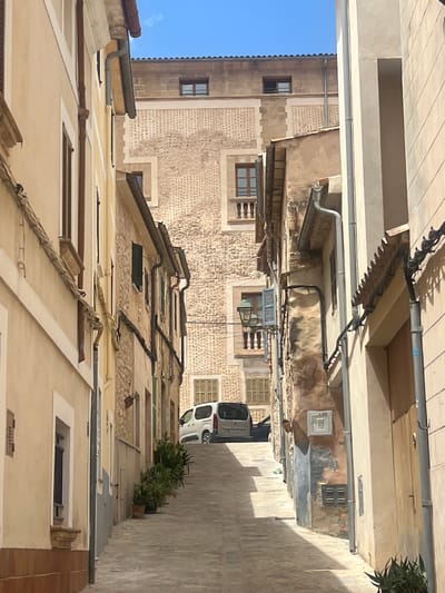 The image depicts a narrow street in the town of Pollença, Spain, with buildings on either side. The street slopes upwards and curves slightly to the left. On the left side of the street, a pale yellow building with a brown trim and a dark-shuttered window is visible. Further up the street, a building with stone accents and a patterned facade is prominent. On the right side, a beige building with a dark tiled roof and a wooden door is in the foreground. A white van is parked on the uphill side of the street. Potted plants line the right side of the street. The sky is clear and blue.