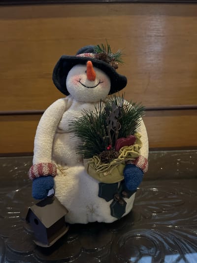 A stuffed snowman decoration is positioned against a wooden background. The snowman is wearing a black hat with red and white trim, a red carrot nose, and has rosy cheeks and a black stitched smile. Its arms are adorned with knitted cuffs, and it is holding a bundle of pine branches, pinecones, and red berries in a burlap-like sack. The snowman also has a small dark brown birdhouse attached to its left side. The background appears to be a paneled wooden wall.