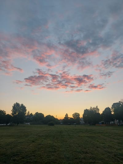 The image shows a wide, grassy field with trees lining the horizon. The sky is a gradient of pale blue at the top, transitioning to a warm orange near the horizon. Wispy clouds are scattered across the sky, tinged with pink and gray from the sunset. In the distance, beyond the trees, some buildings are visible, suggesting a residential area or small town. The field itself appears to be a park or open space, with mowed grass that shows some wear. The overall impression is of a peaceful, late-evening scene in a suburban or semi-rural setting. The location is specified as Strazhitsa, Bulgaria.