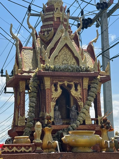 This is a medium shot, eye-level, outdoor photograph of a small, ornate spirit house, likely a San Phra Phum, in Tambon Phraek Ha, Thailand. The spirit house is elaborately decorated with red and gold accents, featuring tiered roofs, intricate carvings, and what appear to be miniature statues of deities or guardians within its central chamber. Garlands of light-colored artificial flowers hang around the structure. The spirit house is situated on a pedestal adorned with golden vases, a lion-like statue, and other decorative items, some of which are weathered or showing signs of age. In the background, a utility pole with numerous black insulators and a complex network of overhead power lines crisscross against a bright blue sky with some scattered clouds. The overall impression is one of a roadside shrine, common in Thai culture, set against a backdrop of urban infrastructure. There are no people visible in the frame.