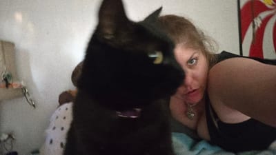 A woman with fair skin, blonde hair, and blue eyes is angled towards the camera, wearing a black top and a necklace with a skull pendant. She is partially lying down on a blue surface. A black cat with yellow eyes, wearing a purple collar, is prominently positioned in the foreground, partially obscuring the view. A white wall, a bedside table with keys, and a poster with red and white stripes are visible in the background.