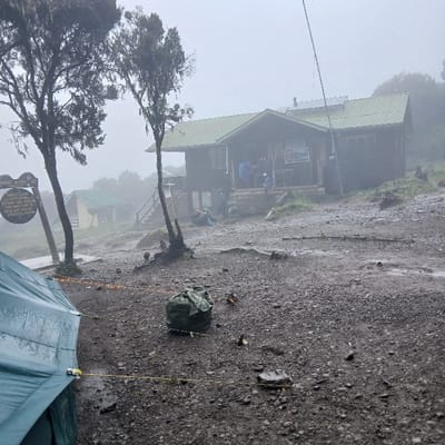 The scene is captured at Horombo Huts, Tanzania, within the implied context of a "city of Horombo Huts" in an elevated, misty environment. The sky is overcast and foggy, obscuring distant features. The ground is dark, wet, and covered in gravel and small rocks, indicative of recent precipitation.

In the immediate foreground, a portion of a teal-green tent is visible on the left, secured by yellow guy lines. To its right, a large, dark green, seemingly packed duffel bag rests on the ground.

In the mid-ground, two slender trees with sparse, moss-like foliage stand, flanking a dark wooden signpost with white lettering. Behind these, a two-story wooden hut with a green corrugated metal roof is elevated on a stone or concrete foundation. The hut features a front porch with stairs. On the roof, two solar panels are visible, along with a tall pole extending upwards, possibly an antenna or flagpole.

Several individuals are present on and around the main hut. On the front porch, multiple figures are visible; one person wears a blue jacket, and a smaller figure is seated on the steps. Other indistinct individuals are inside the porch area. Another person is discernible near the base of the hut on the left, and a dark-clad figure sits on the ground towards the right side of the hut.

Further into the background, partially obscured by mist and sparse vegetation, are at least two smaller structures, appearing to be additional huts, also featuring green roofs. The overall scene depicts a functional mountain camp or waypoint with active human presence.