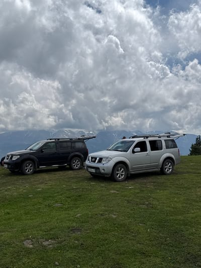 Two Nissan Pathfinder SUVs are parked on a grassy incline in a mountainous region. The vehicle on the left is black, and the vehicle on the right is silver. Both vehicles have their rear hatches open. The SUVs are facing towards the right of the frame. In the background, snow-capped mountains are visible under a cloudy sky. A single conifer tree is present to the far right of the frame. The location is identified as Nizhnyaya Teberda, Russia.