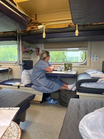 The image depicts the interior of a train compartment, likely in the city of Kursk, Russia, given the provided context. A woman with blonde hair, wearing glasses and a blue and white striped dress, is seated at a small table by the window. She is looking out at the passing scenery. The compartment features bunk beds, characteristic of train travel, with some beds made up with bedding. A dark green duffel bag is on the floor next to the woman, and she is wearing blue Crocs. Various items are on the table, including what appears to be tea or coffee packets and a small bottle. The overall environment suggests a journey in progress.