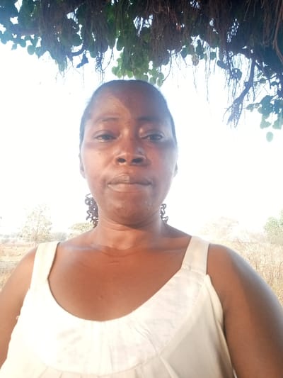 A woman with dark skin and short hair stands beneath a leafy tree canopy. She is wearing a white sleeveless top. Her expression is neutral, and she is looking directly at the camera. A landscape with trees and a clear sky stretches out behind her, although the background is somewhat washed out by the bright light.