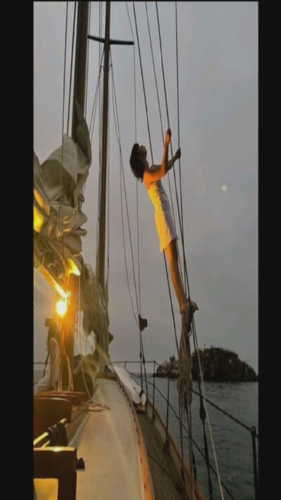 A woman in a white dress is climbing the rigging of a sailboat. She is high up on the boat and holding onto the ropes. The boat is on the water, and there is an island in the background. The sky is overcast and the sun is setting, creating a golden light. The sails are furled and the deck is visible.
