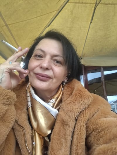 A woman poses under a yellow awning, cigarette in hand. She wears a brown fur-like jacket over a gold and white scarf. Her dark hair frames a face with a soft, neutral expression. A ring adorns her finger, and dangling earrings are visible. Behind her, a window reflects an outdoor scene, suggesting a cafe or restaurant setting.