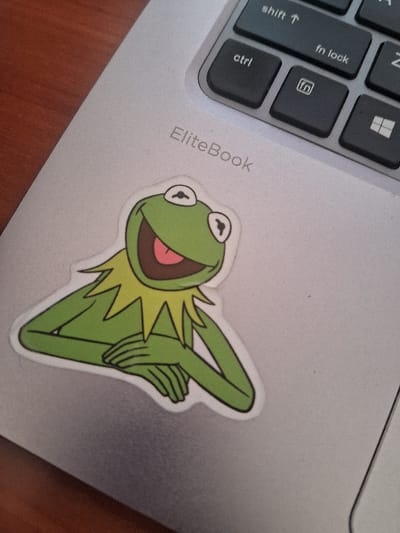 A close-up, overhead image depicts a silver-colored HP EliteBook laptop resting on a reddish-brown surface, likely a wooden table, in the general context of Muthambi ward, Kenya. A sticker of Kermit the Frog is affixed to the laptop's palm rest area. The sticker shows Kermit from the waist up, facing slightly right, with a wide open-mouthed smile revealing a pink tongue, large white eyes with black pupils, a green body, and a prominent yellow pointed collar. His hands are clasped in front of him. The sticker has a distinct white outline. A portion of the laptop's black keyboard is visible in the upper right quadrant, displaying keys such as "ctrl", "shift ↑", "fn lock", a key with a Windows logo, and another with a LinkedIn logo. The "EliteBook" branding is embossed on the laptop surface above the sticker.