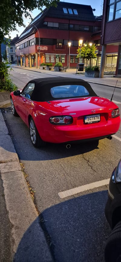 In Lillehammer, Norway, a red Mazda convertible is parked on the side of a street. The building behind the car displays the word "POLITI" in neon lights, indicating it is a police station. The car is parked on asphalt, adjacent to a concrete curb. Streetlights are illuminated, suggesting it is evening or early morning. The environment appears to be a typical urban street scene with buildings and a road.