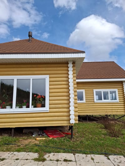 This image captures a vibrant yellow house with a brown shingled roof under a bright blue sky adorned with white cumulus clouds. The house features a distinct log cabin-style exterior, with vertical yellow planks and white decorative trim, including a scalloped border above the lower windows.
Multiple white-framed windows are visible on the house's facade. The largest window on the ground floor reflects potted plants, predominantly red and pink geraniums. Above it, a smaller window is present, and another multi-paneled window is seen on the upper story to the right. A dark vent pipe protrudes from the brown, hexagonally shingled roof.
The surrounding environment includes a sloped grassy area to the right, showing various shades of green and some dry patches. A pathway of light-colored paving stones, with grass growing in the seams, runs along the far right edge of the frame. A black pipe extends along the ground near the house. Behind the building, parts of bare trees or bushes are visible. On the ground next to the house, a red object with the text "Coca-Cola" written on it multiple times, possibly a cooler or storage box, is discernible, along with a yellow cylindrical object.
The scene depicts a peaceful daytime setting, likely a residential property or dacha in Ludorvai, Russia. No people or specific activities are present, focusing instead on the architectural details of the house and its immediate surroundings under clear weather conditions.